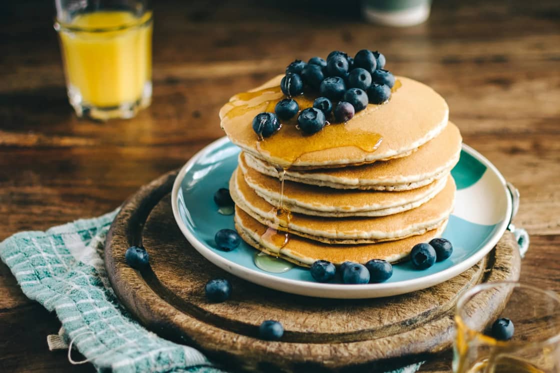 A tall stack of pancakes topped with blueberries on a blue plate, with orange juice, a syrup jar and a fork on a dark wooden table