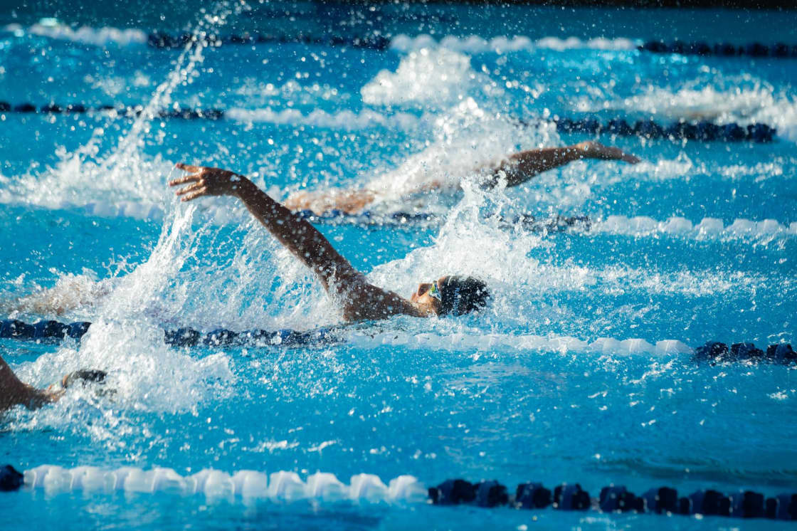 Two swimmers racing freestyle in neighboring lanes of a sunlit outdoor pool, one arm reaching forward through heavy white spray between blue-and-white lane lines