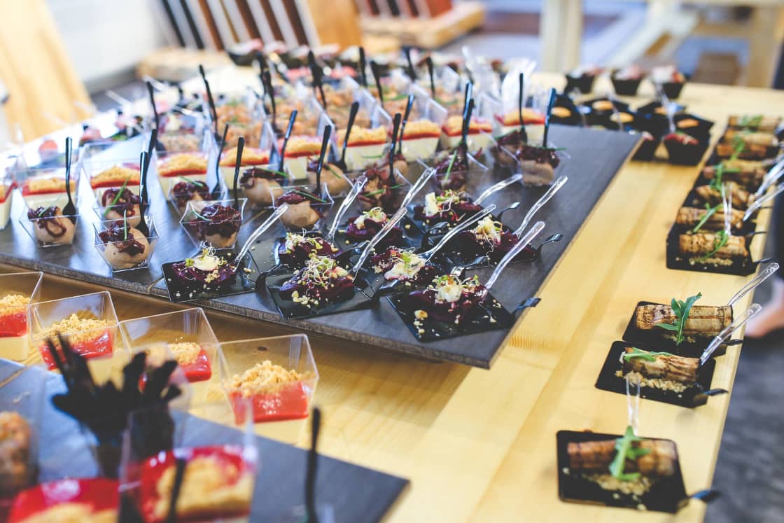 Rows of small tasting portions laid out on a wooden table: dark beetroot pieces under a scatter of micro-greens on black square dishes, clear cups of layered dessert showing a red layer beneath a crumb topping, and glazed rolls under herb garnish, a clear plastic fork or spoon stood in every one