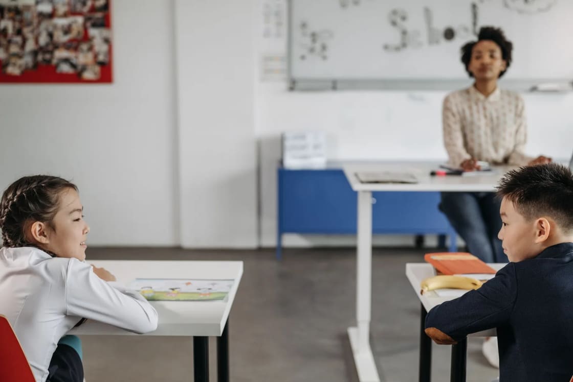 A teacher standing at the back of a classroom while two young pupils work at desks in the foreground beside a whiteboard