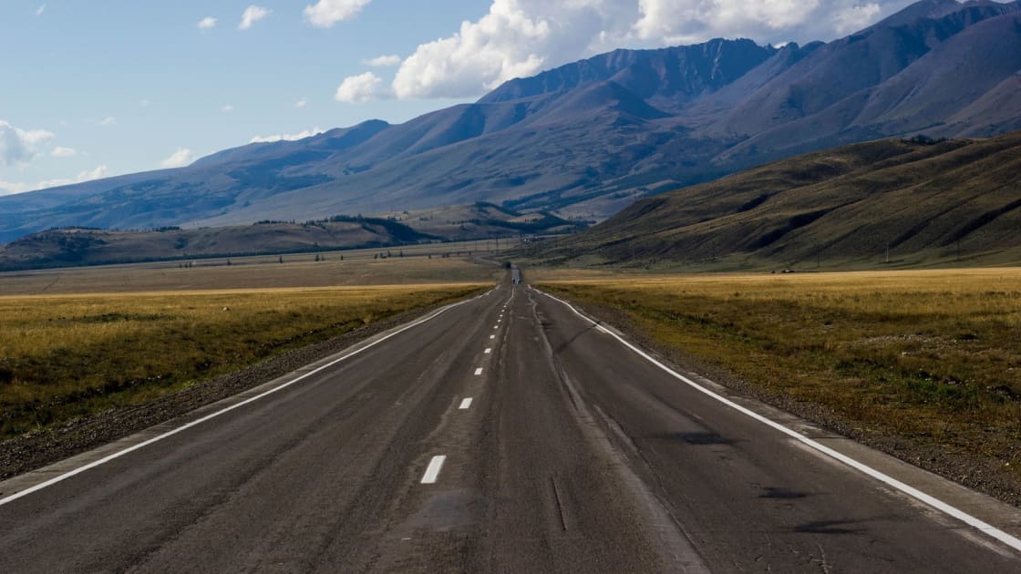 A straight two-lane highway stretching across open grassland toward a long mountain range under a blue sky