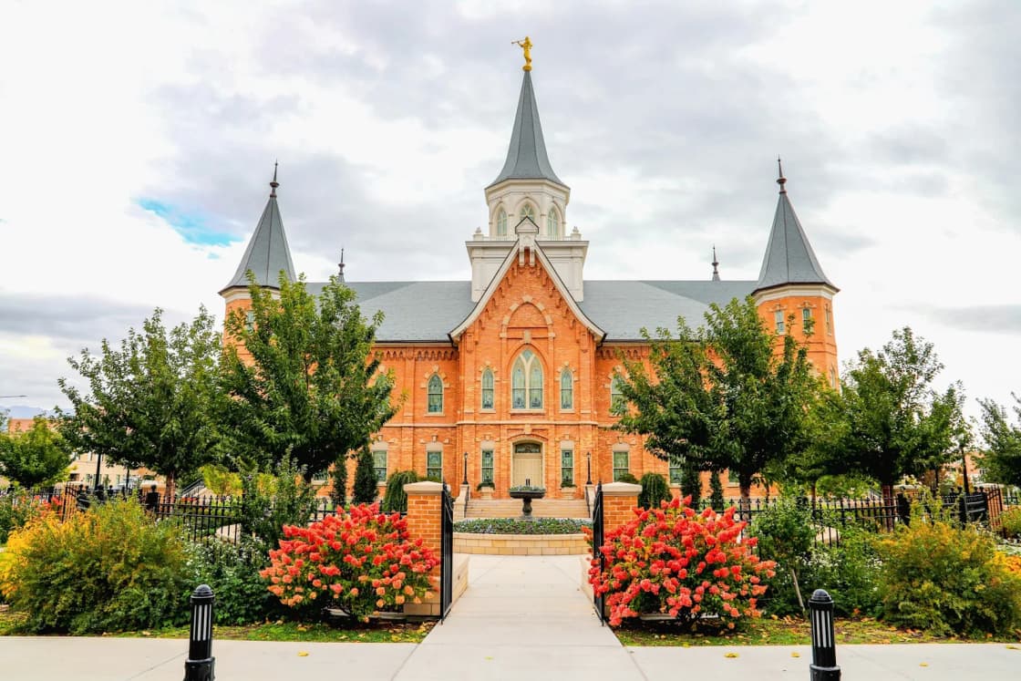 The red-brick Provo City Center Temple with its gray spires and gold statue, seen through the front gate past beds of red flowers and trees.