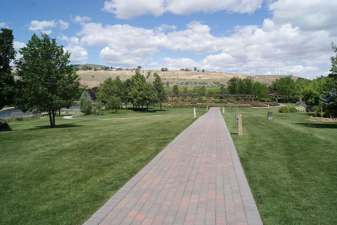 A brick path crosses green lawns toward the rose garden arbor at Thanksgiving Point, with dry hills behind.