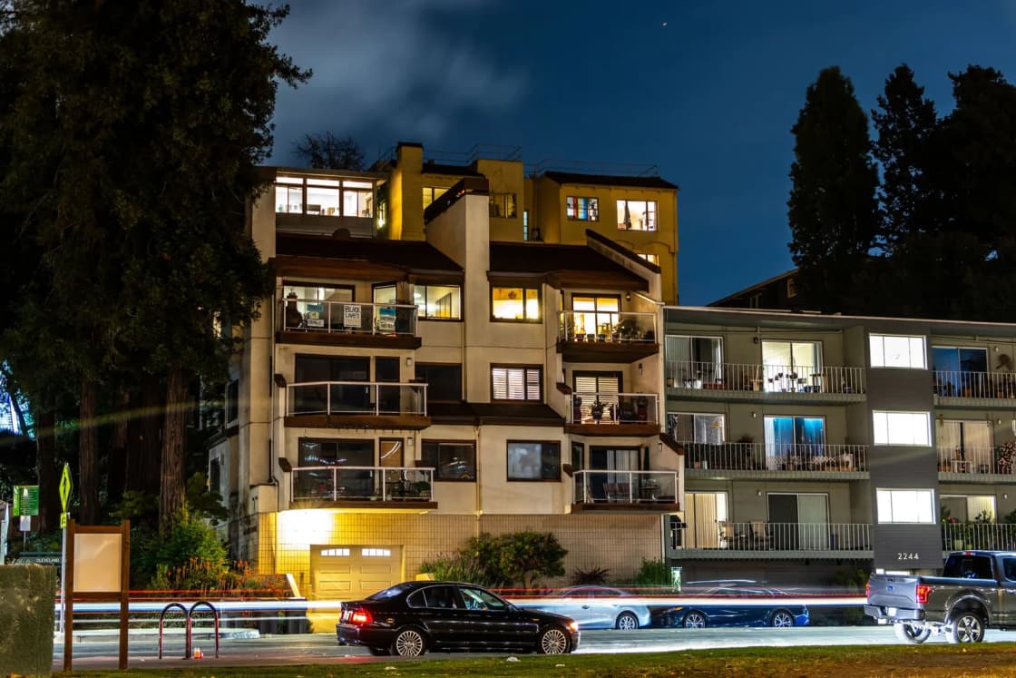 A low-rise apartment building with balconies and lit windows at dusk, with cars parked along the street.