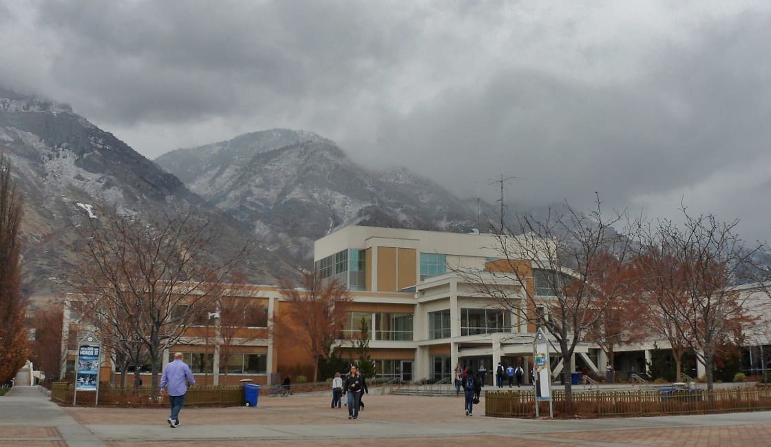 The Wilkinson Student Center at BYU, a tan and glass building beyond a brick plaza where students are walking, with bare trees and snow-dusted mountains under low clouds