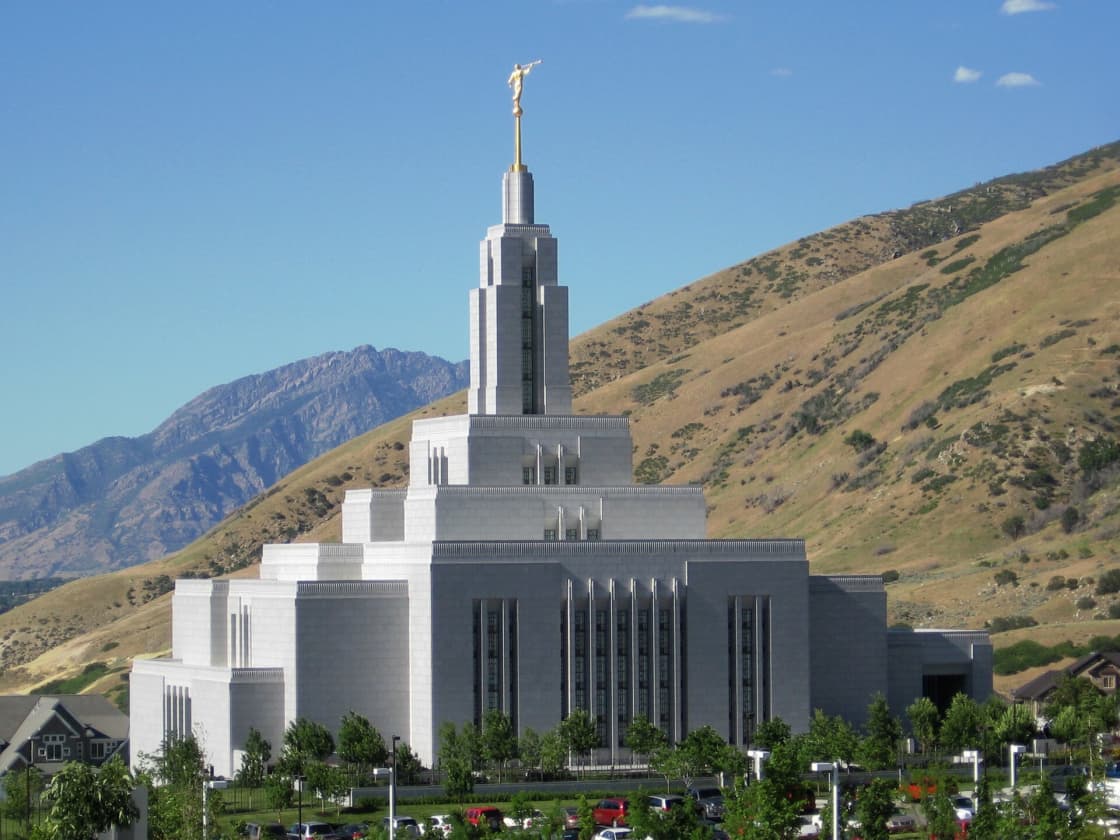 The white granite Draper Utah Temple with its tiered spire and gold angel Moroni statue against a golden foothill and a rocky peak under a blue sky, with trees and a parking lot in front