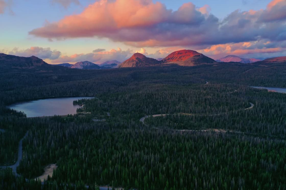 An aerial view of dark forest and small lakes along the Uinta front above Kamas at sunset, with pink clouds over the peaks.