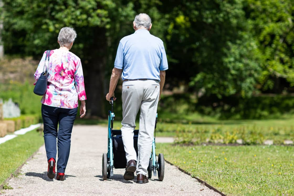 An older couple seen from behind walking along a sunny gravel park path between lawns and trees, one in a floral blouse and one in a blue shirt pushing a rollator