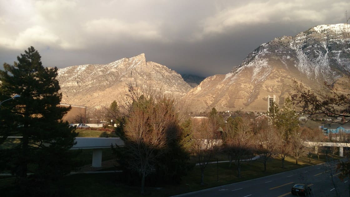 Snow-dusted peaks at the mouth of Rock Canyon under dark storm clouds, seen from BYU's campus, with the Centennial Carillon Tower, bare trees, a footbridge and a road below and no snow on the valley floor