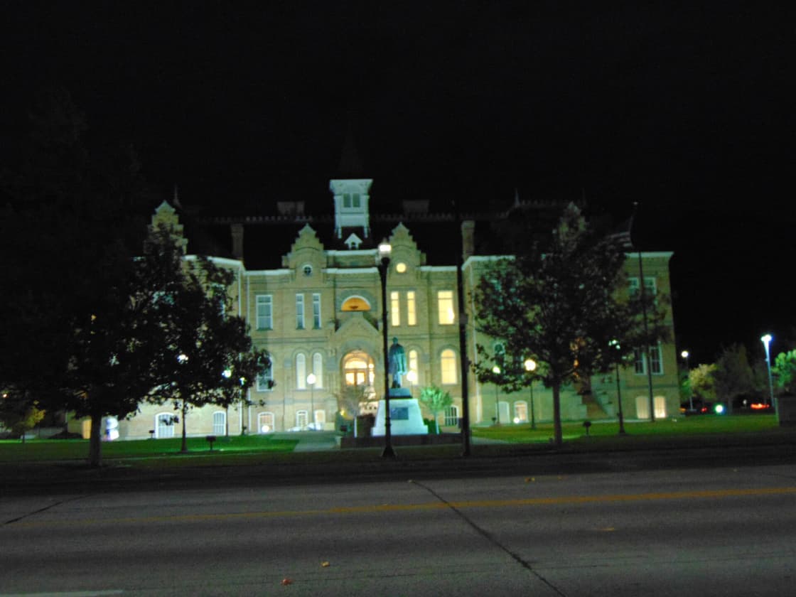 The historic Provo City Library building at Academy Square lit up at night, seen across University Avenue, with a statue on a pedestal and trees on the front lawn