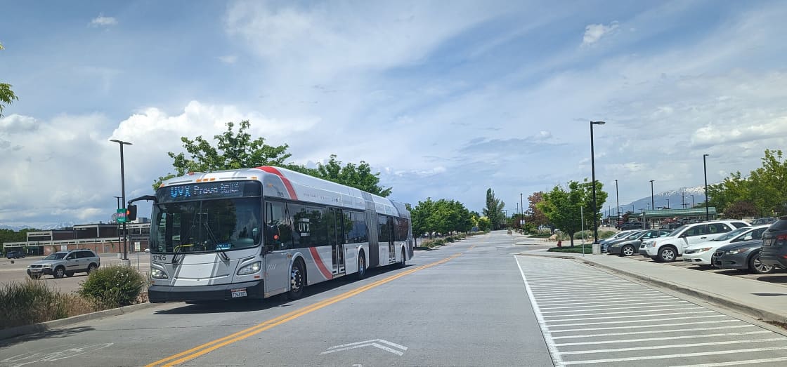 A silver articulated UVX bus with a red stripe and a Provo destination sign on the road beside the park-and-ride lot at Orem Central Station, with snow-capped mountains in the distance