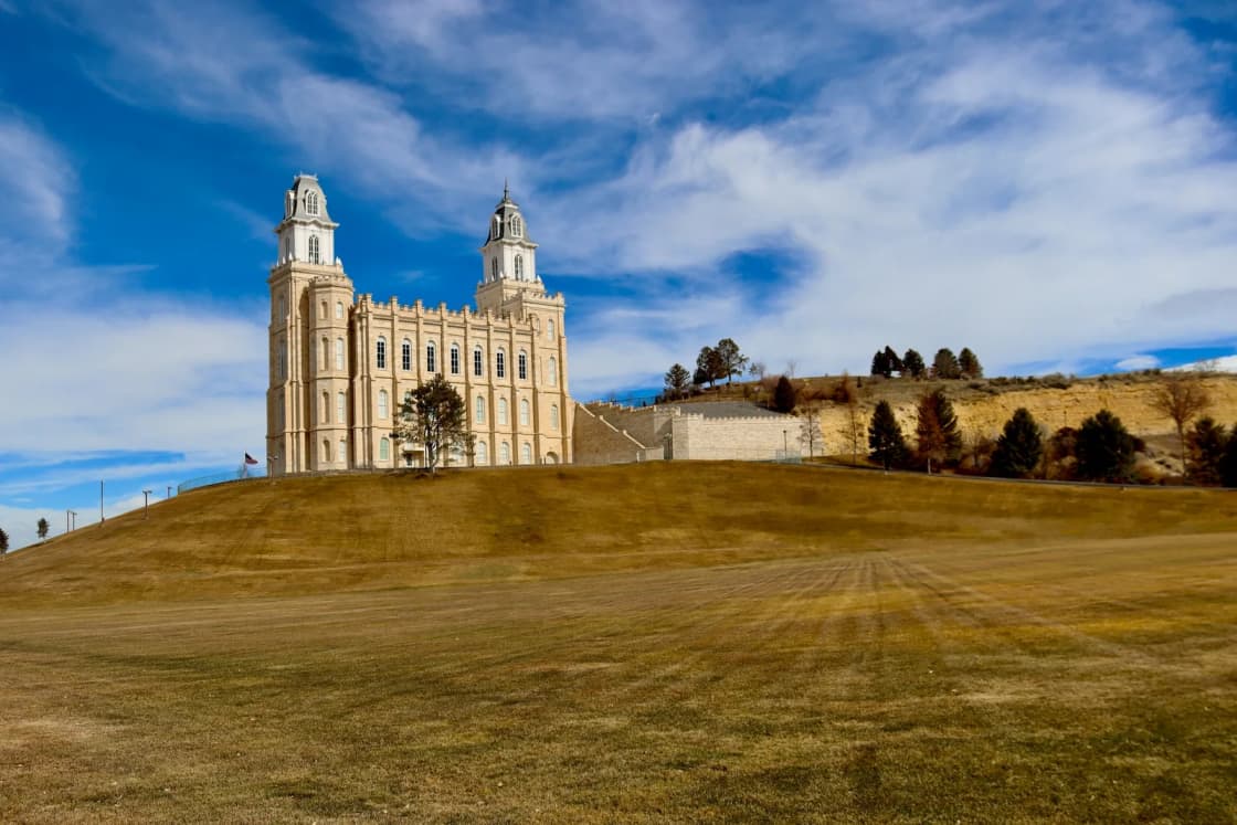 The cream limestone Manti Utah Temple with its twin towers stands atop its grassy hill under a blue sky streaked with white clouds.