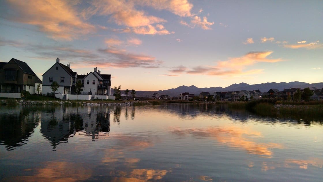 Sunset over Oquirrh Lake in Daybreak, with pink and orange clouds reflected in the still water, lakeside homes along the shore and mountains on the horizon