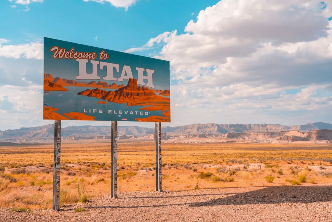 A Welcome to Utah roadside sign standing in open desert scrub, red rock mesas and a big cloudy sky behind it