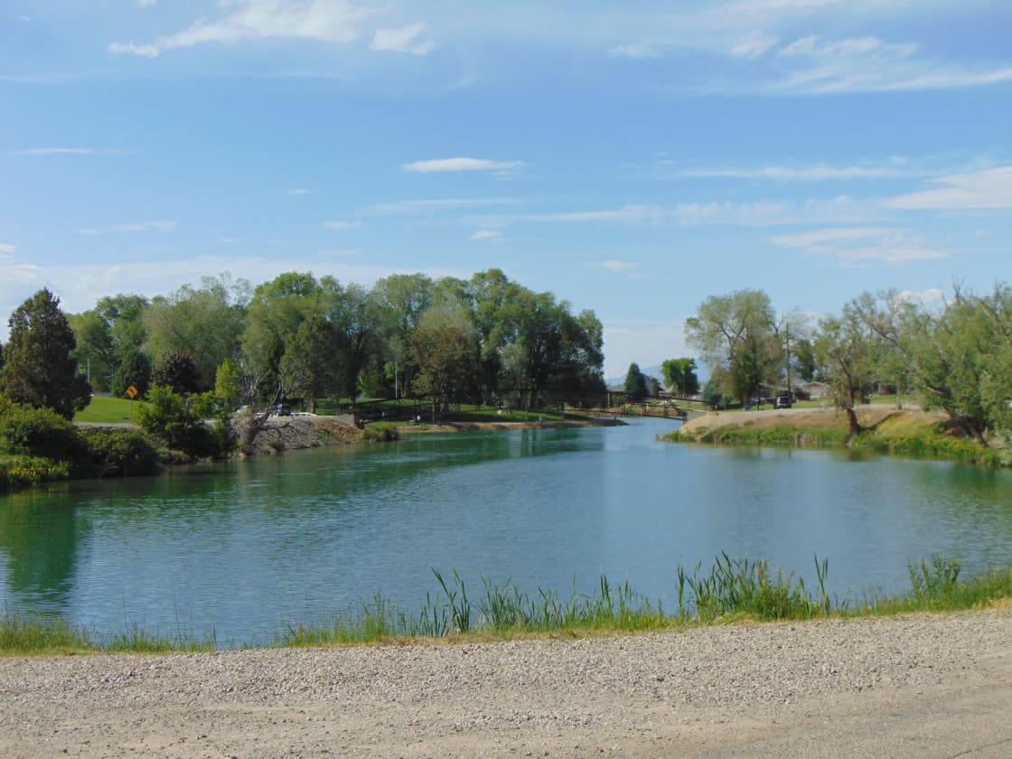 Blue-green pond with a footbridge and park lawn on the far bank under a partly cloudy sky