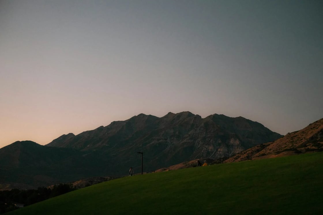 The Wasatch peaks above Provo dark against a fading pink and gray twilight sky, a lone walker on a grassy hillside below