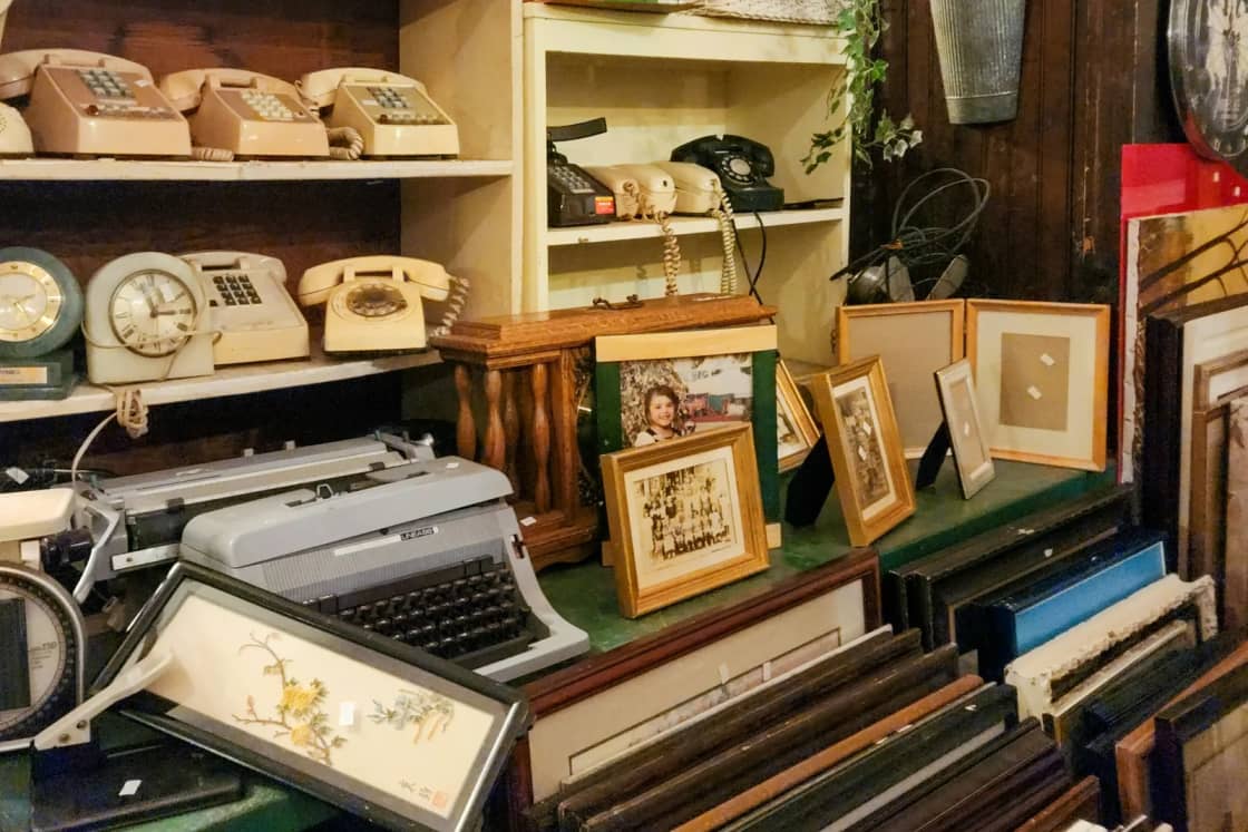 Shelves in a secondhand shop crowded with old rotary phones, a typewriter, clocks and stacks of empty picture frames waiting to be reused