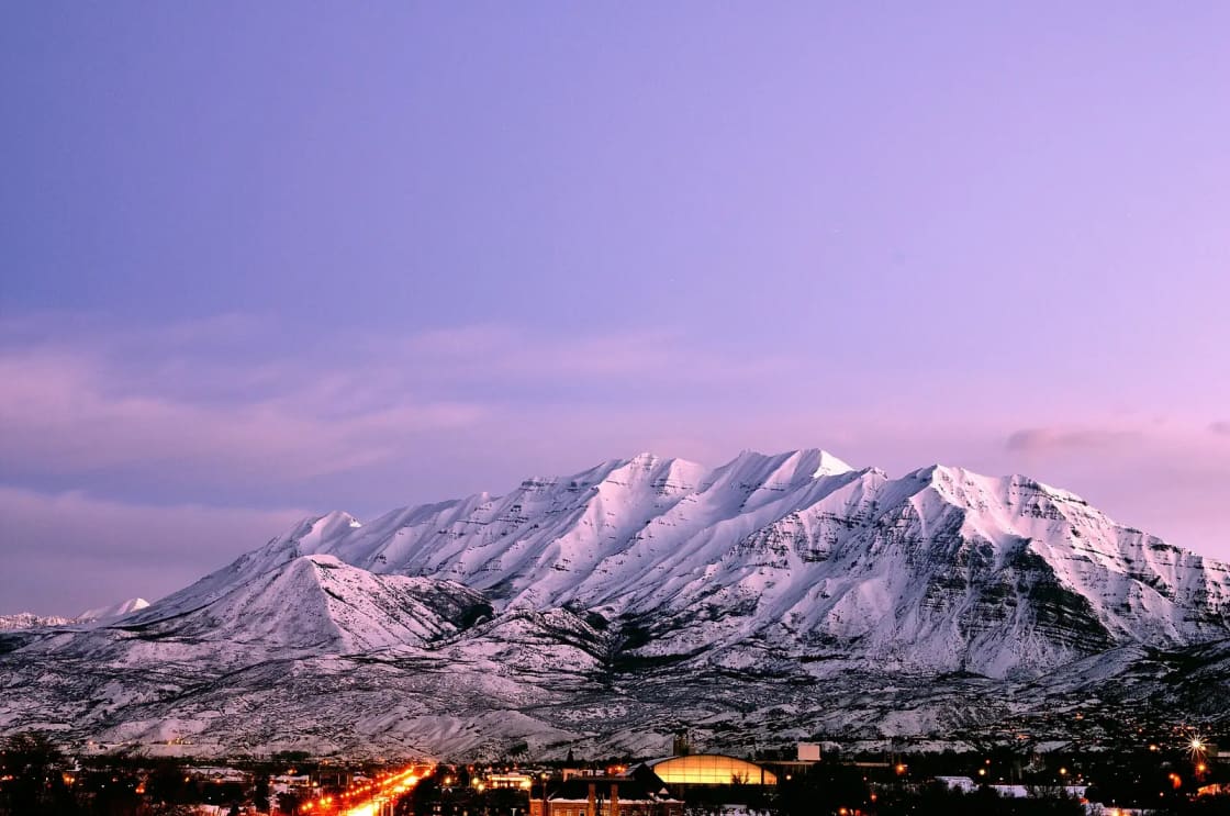 The long snow-covered ridgeline of Mount Timpanogos at dusk under a violet sky, with the lights of the valley strung along its base