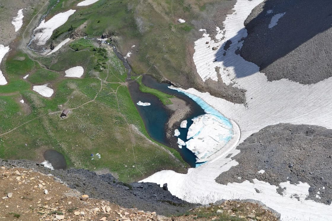 Looking down from the summit ridge of Mount Timpanogos at Emerald Lake, half covered in ice, with snowfields and green alpine meadows in the basin below