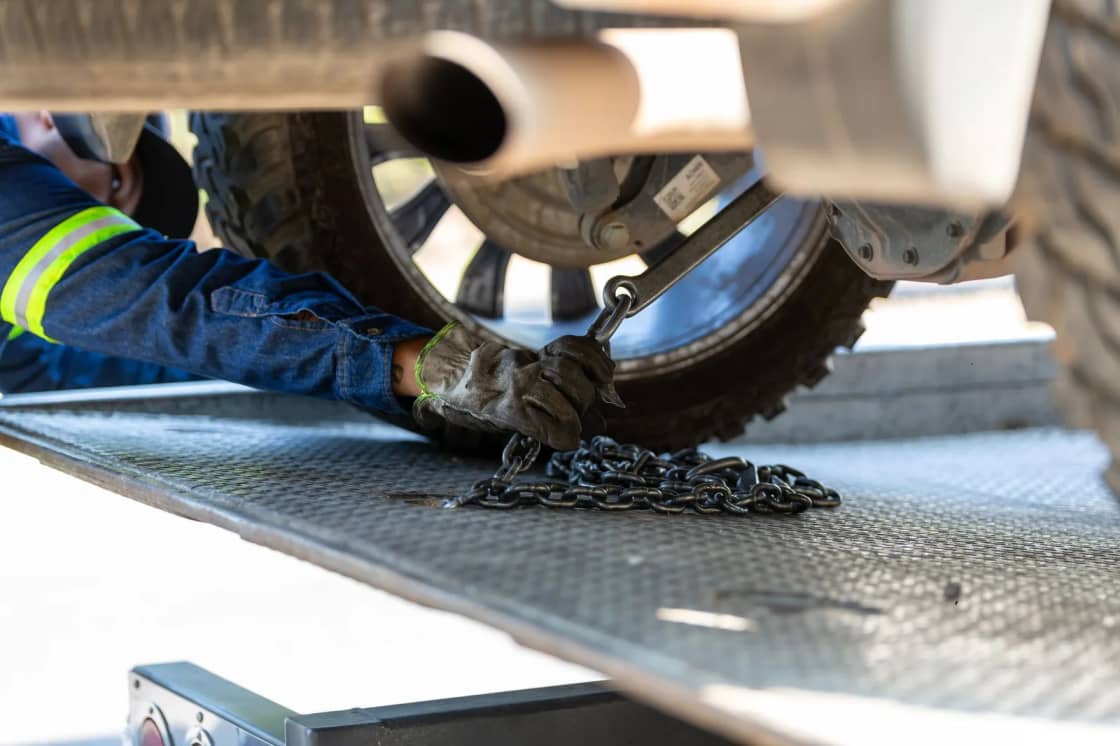 A worker in high-visibility clothing securing a chain around a vehicle wheel on a flatbed tow truck deck