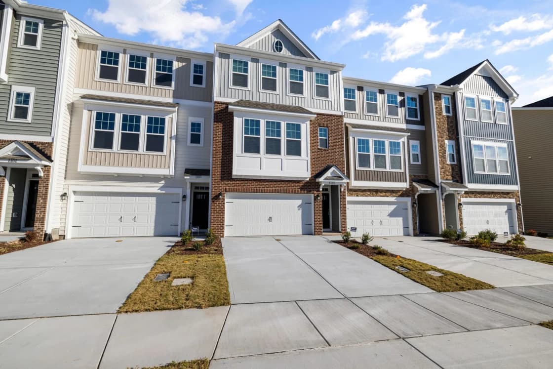 A row of newly built three-story townhomes with attached garages, mixed brick and siding fronts, and concrete driveways running to the sidewalk under a blue sky
