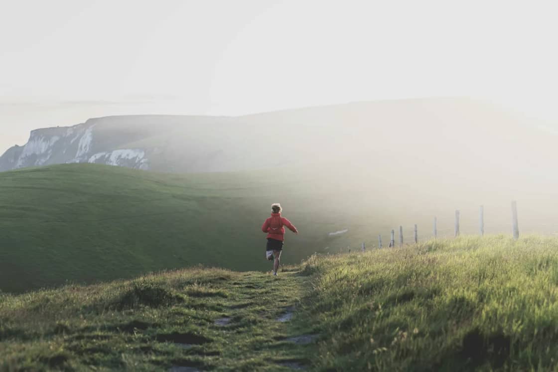 A lone runner in red heading up a narrow dirt path across a grassy hillside, with cliffs fading into morning fog behind