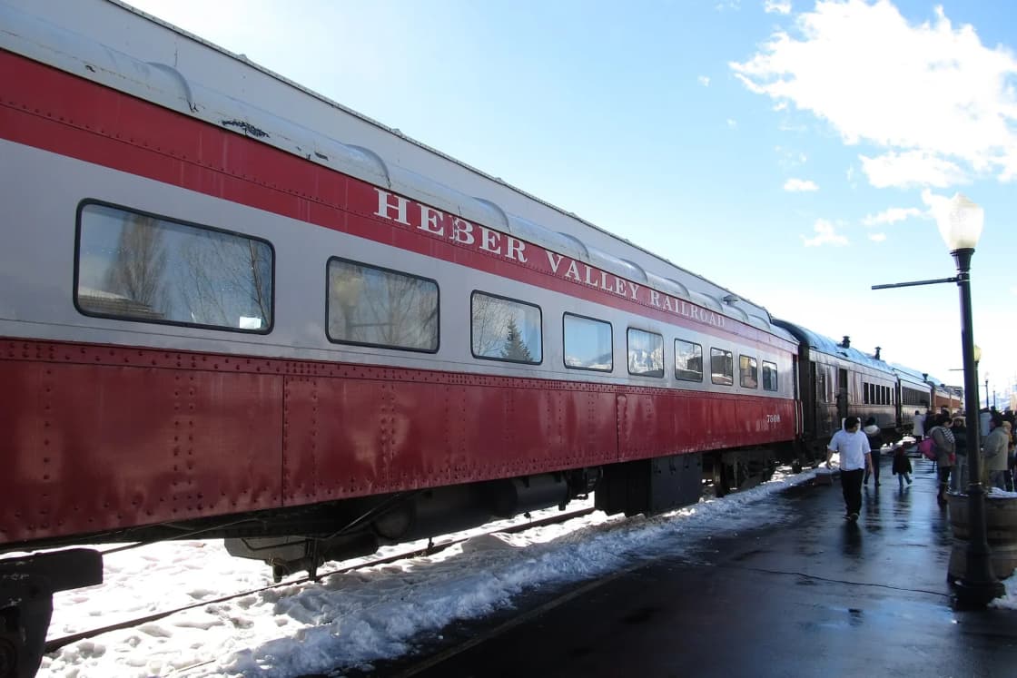 A red and silver Heber Valley Railroad passenger car waits at a snowy platform as riders walk alongside.
