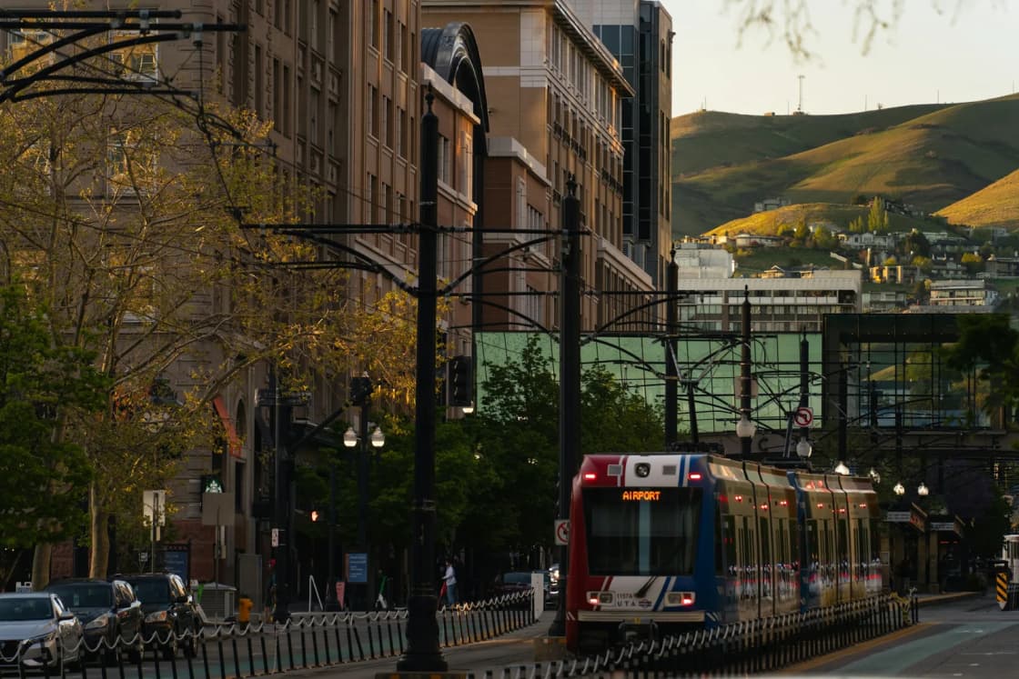 A red, white and blue TRAX light-rail train marked Airport heads down a tree-lined downtown Salt Lake City street toward the foothills.