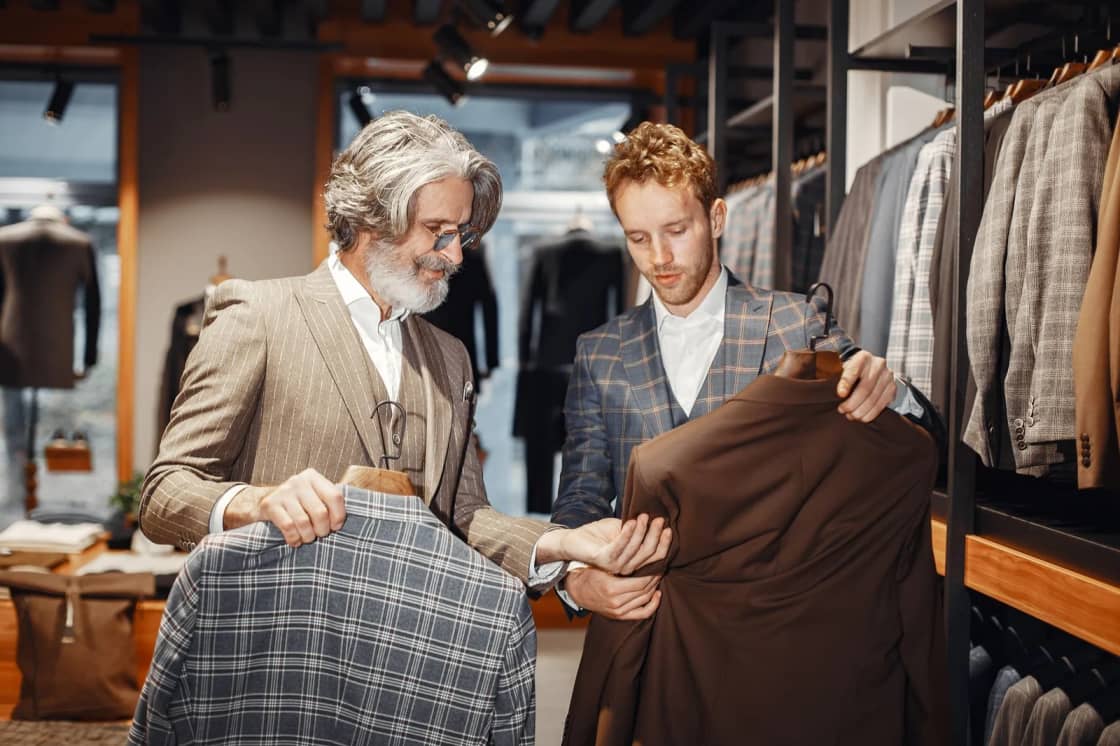 Two men in a menswear shop comparing jackets on hangers in front of racks of suits