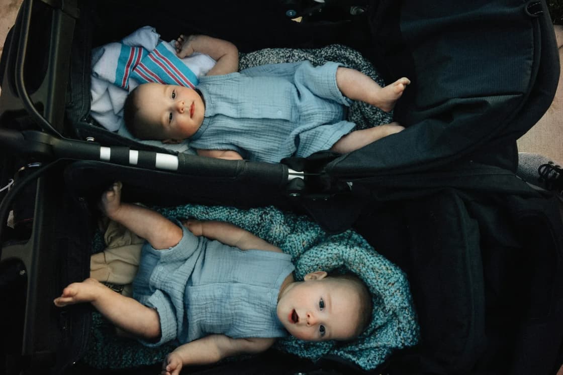 Twin babies in matching blue rompers lying side by side in the two seats of a double stroller, seen from above.