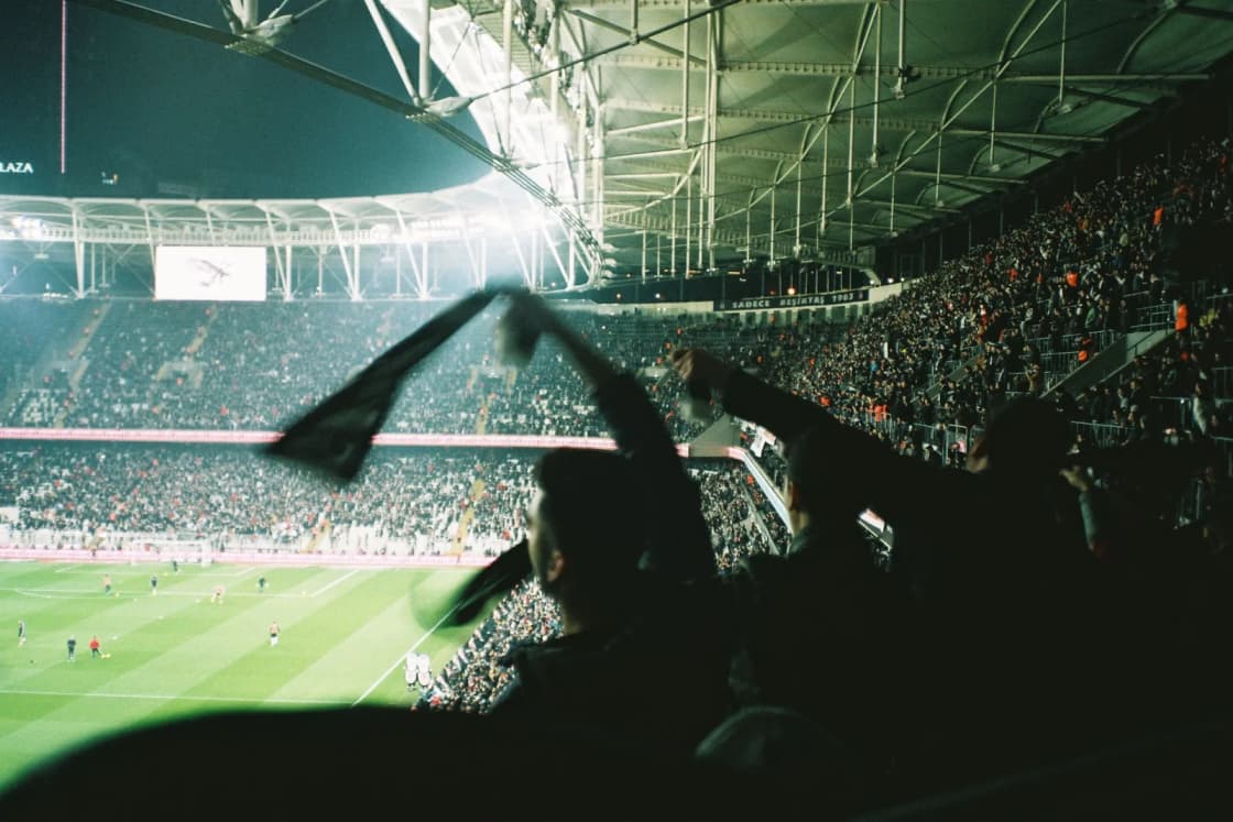 Fans in silhouette waving scarves above their heads in a packed stadium under bright lights at night