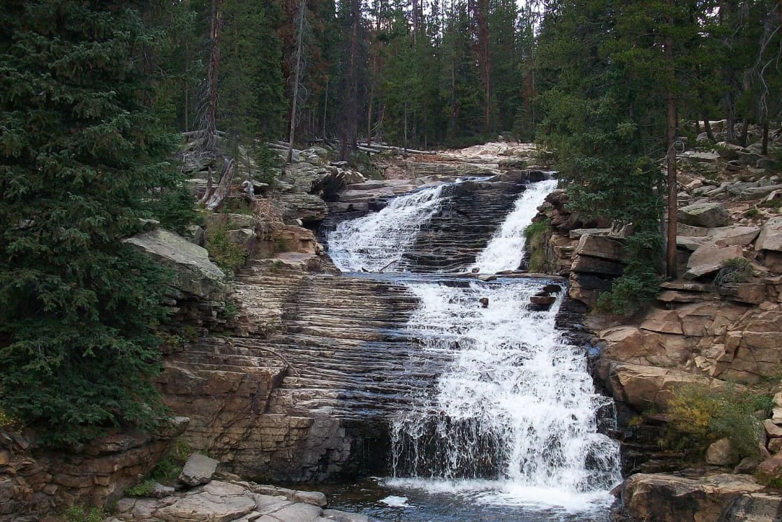 The Upper Provo River tumbles over layered rock ledges in a cascade surrounded by dark evergreen forest.