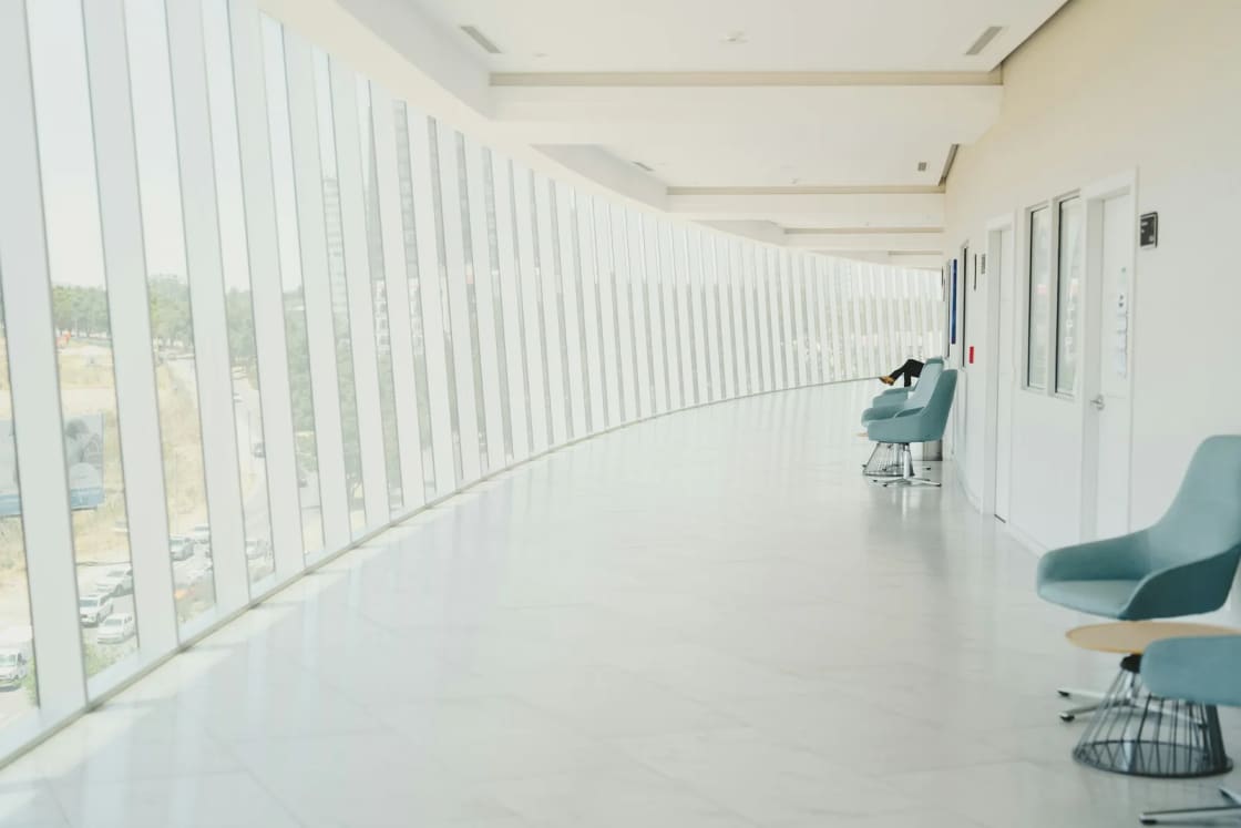 A long, bright clinic corridor lined with floor-to-ceiling windows on one side and a few teal waiting chairs along the wall