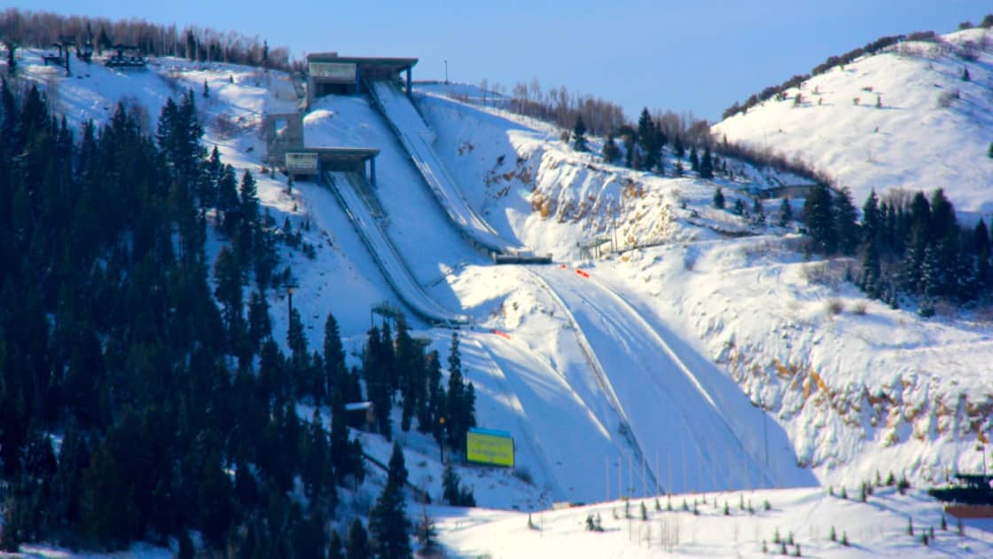 The Utah Olympic Park ski jumps near Park City in winter, their inrun towers at the top of a long snow-covered landing hill between dark evergreens and white slopes under a blue sky