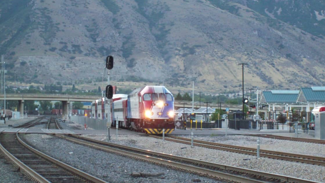 A FrontRunner train leaving Provo station with its headlights on, the tracks and signals in front and the station's teal canopies and the Wasatch mountainside behind