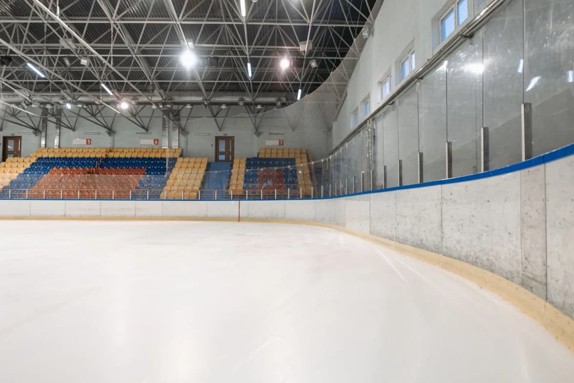 An empty indoor ice rink, freshly resurfaced, with dasher boards, glass, and rows of blue, yellow, and red bleacher seats under bright lights — Peaks Ice Arena's role in Utah 2034