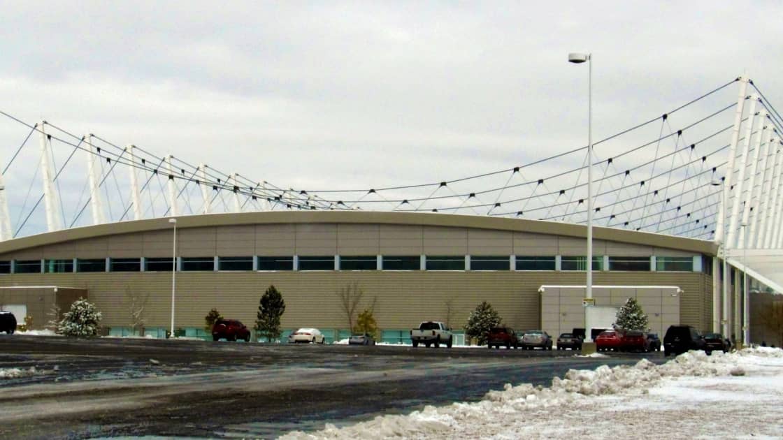 The Utah Olympic Oval in Kearns on a snowy winter day, its long low building hung from a row of white masts and cables, with cars parked out front and snow piled along the lot