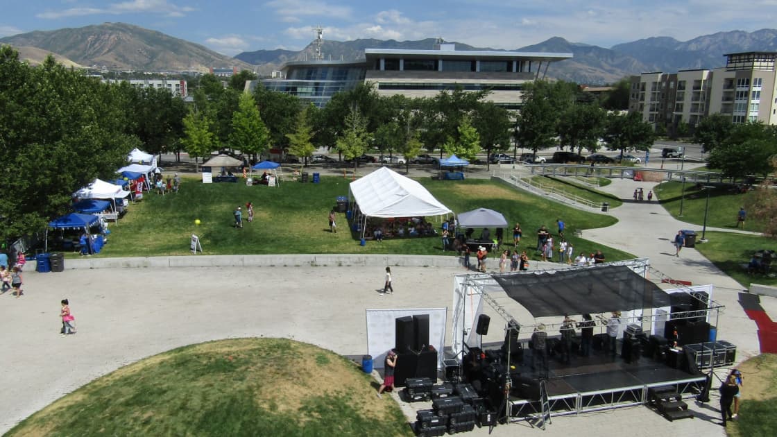 An outdoor stage and white and blue event tents on the lawns of Library Square in downtown Salt Lake City, seen from above, with the library, apartment towers and the Wasatch Mountains beyond