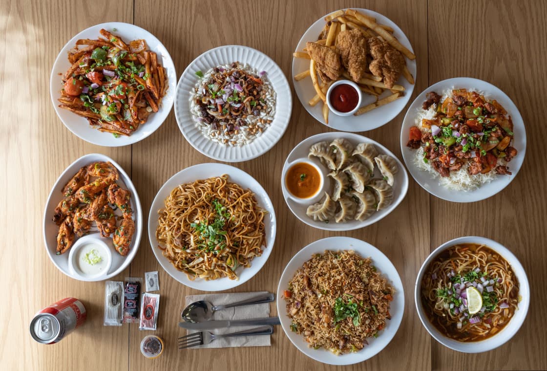 An overhead spread of dishes on a light wood table: stir-fried strips of meat and peppers, a puffed-rice salad, breaded chicken and fries beside a pot of ketchup, chili chicken over white rice, spiced wings and a dip, a plate of chow mein, steamed dumplings around an orange dipping sauce, fried rice, and a bowl of noodle soup topped with red onion and a lemon wedge