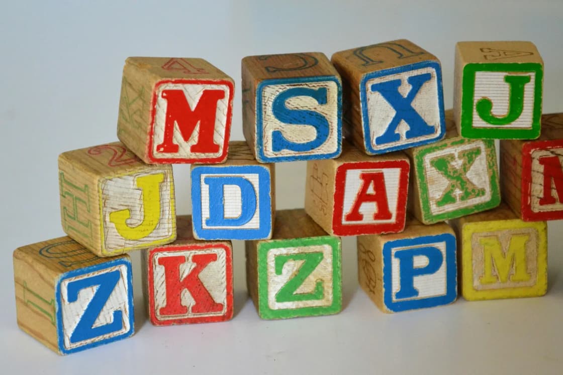A stack of worn wooden alphabet blocks painted with red, blue, green and yellow letters on a pale background.
