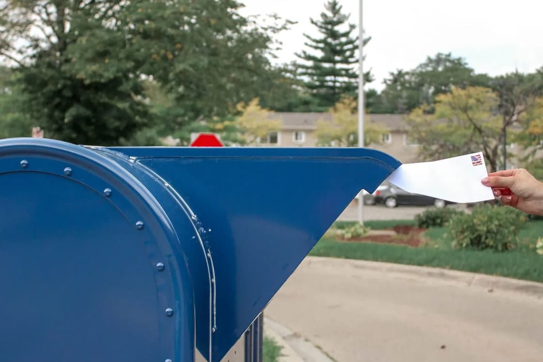 A hand dropping a stamped envelope into a blue curbside mailbox, with an American flag flying on a pole behind it