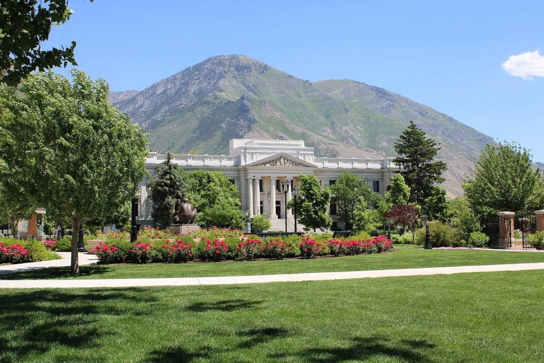 The white columned Historic Utah County Courthouse behind a lawn and red rose beds, with a mountain rising behind it.