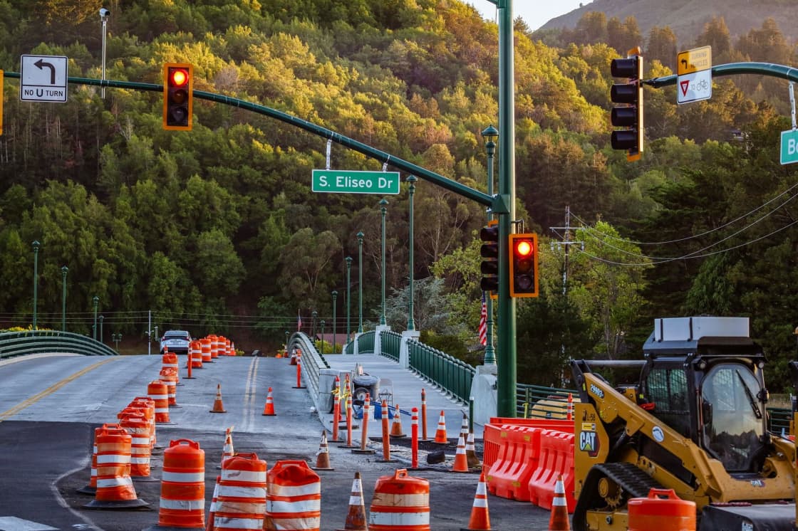 Orange barrels and construction equipment narrowing lanes on a road beneath a traffic signal