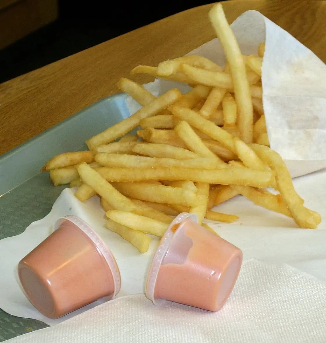 Two small plastic cups of pink fry sauce sitting beside a paper cone of french fries on a diner tray