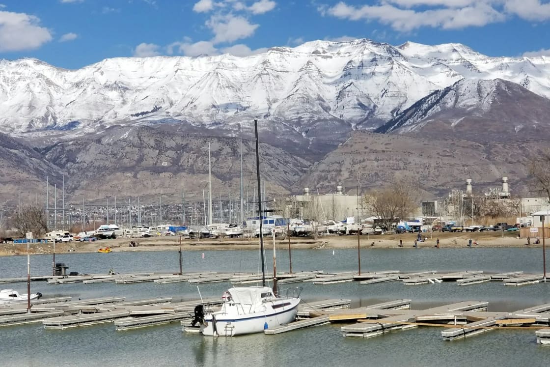 A sailboat tied up among mostly empty slips at a Utah Lake marina on the Vineyard shore, snowy Wasatch peaks behind