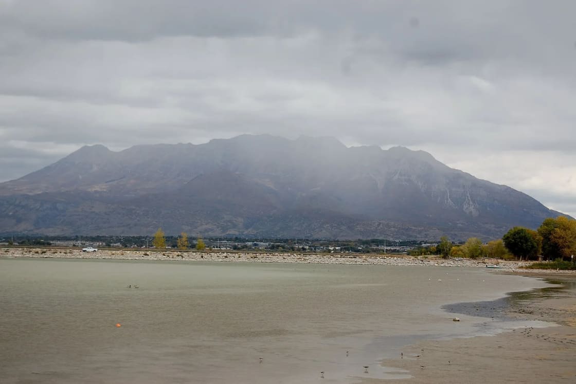 Mount Timpanogos rises under gray clouds beyond the shallow water and shoreline of Utah Lake State Park.