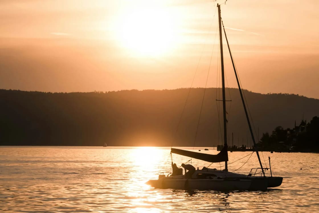 A small sailboat silhouetted on a lake at sunset, the sun low over dark hills and a gold path across the water
