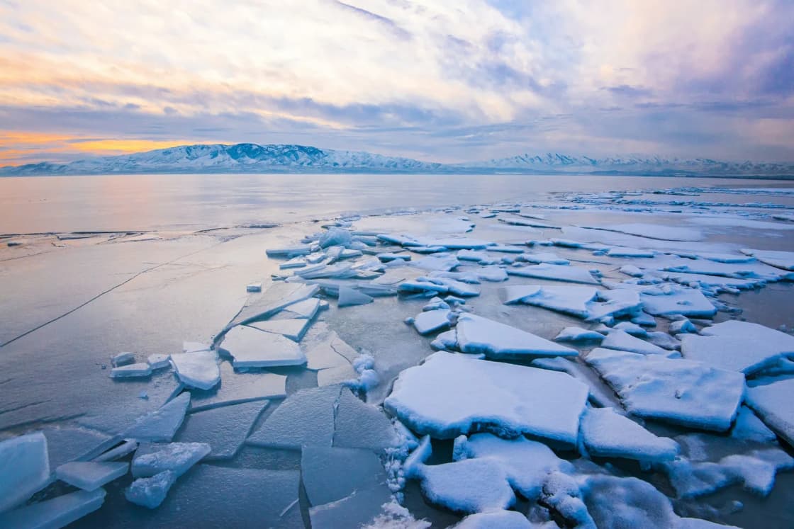 Broken plates of ice piled along the edge of Utah Lake in winter, open water and snowy mountains beyond under a pastel sunset sky