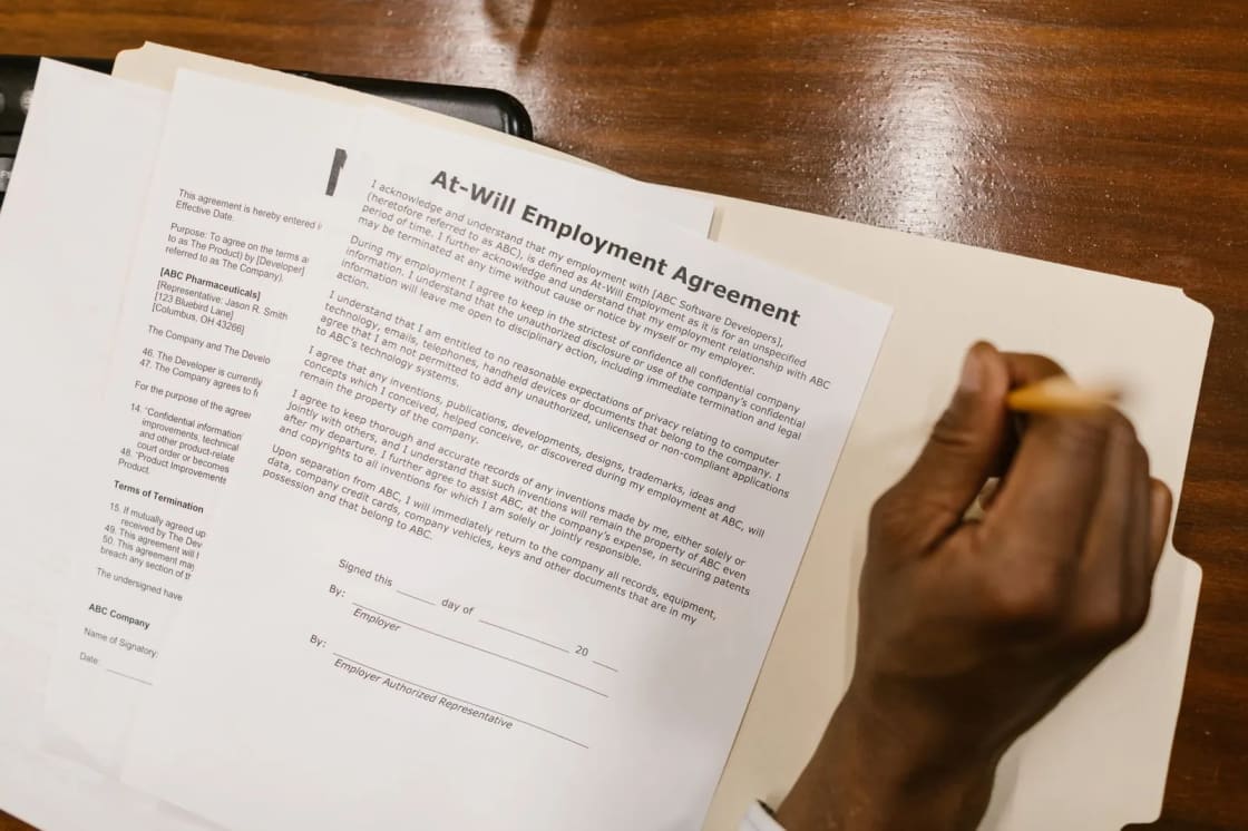 A printed template headed At-Will Employment Agreement lying open in a manila folder on a dark wood desk, its visible clauses covering confidentiality and invention assignment, with a hand holding a pencil beside the signature lines