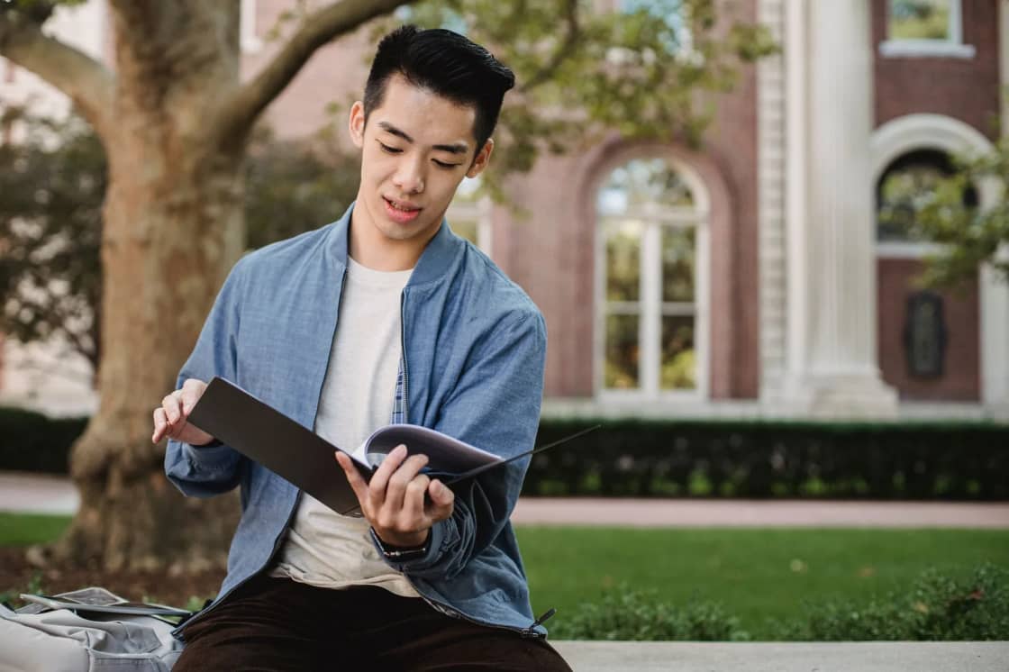 A student in a denim jacket standing outside a brick campus building, reading from an open folder of papers